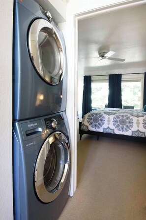 Ein Zimmer mit Waschmaschine, einem Bett mit gemusterten Betttextilien, einem Deckenventilator und einem Fenster mit Vorhängen.