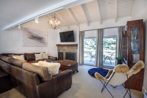 A living room with a sectional sofa, a fireplace, a TV, and a large window with a view. - Snow Creek Comfort -2 Bedroom + Loft -outdoor pool (Sun Valley)
