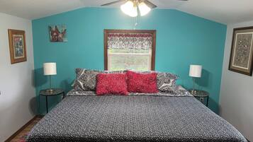 A bedroom with a bed, two bedside tables with lamps, a ceiling fan, a window with a floral curtain, and wall art.