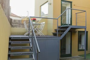 Eine Treppe führt zu einem Balkon mit Metallgeländer, einem Tisch mit Stühlen und einem Fenster mit Blick ins Freie.