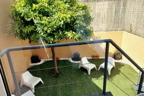 Ein Balkon mit künstlichem Rasen, weißen Plastikstühlen und einem Blumentopf.