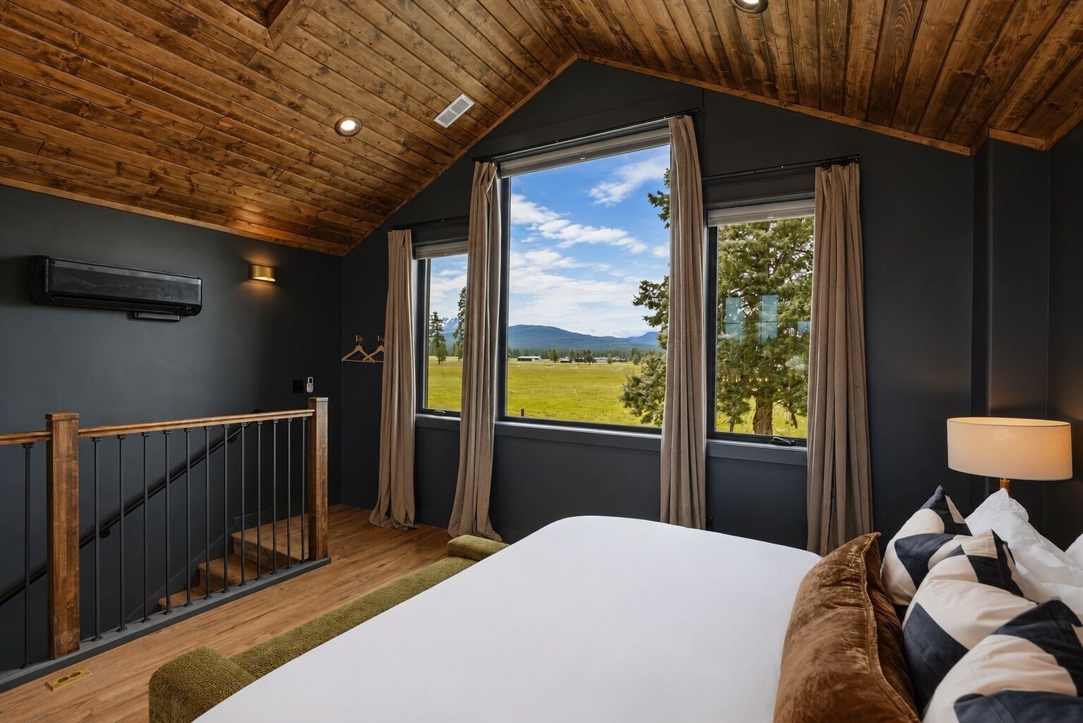 A bedroom with a large bed, a wooden ceiling, a view of a green landscape, and a staircase.