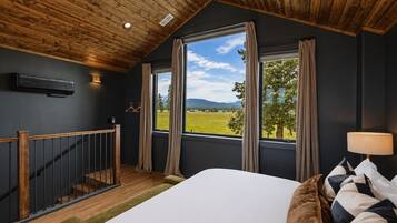 A bedroom with a large bed, a wooden ceiling, a view of a green landscape, and a staircase.