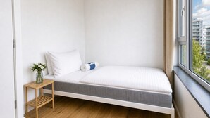 A small, single-bed room with a wooden floor, a bedside table with a vase, and a window with a city view. - Sleek & Modern New Build Flat (Prague)