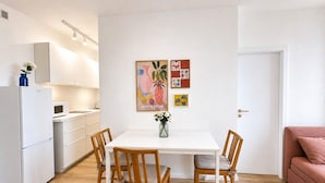A modern kitchen with white cabinets, a white refrigerator, and a dining table with four chairs. There are two framed artworks on the wall above the table. - Sleek & Modern New Build Flat (Prague)