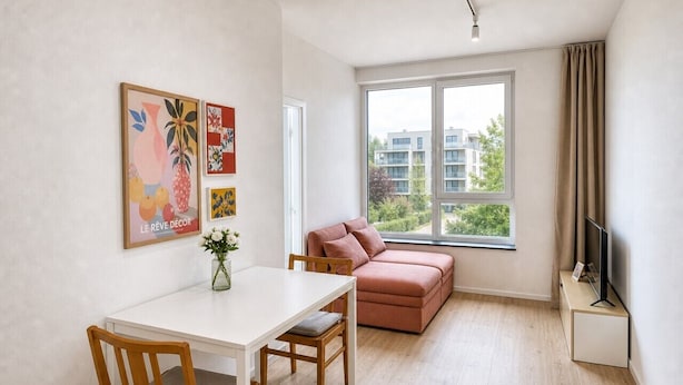 A modern living room with a white table, wooden chairs, a pink sofa, a TV on a stand, and large windows with a view of buildings and greenery. - Sleek & Modern New Build Flat (Prague)