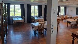Un espace repas comprenant plusieurs tables et chaises, de grandes fenêtres avec des rideaux et un sol carrelé.