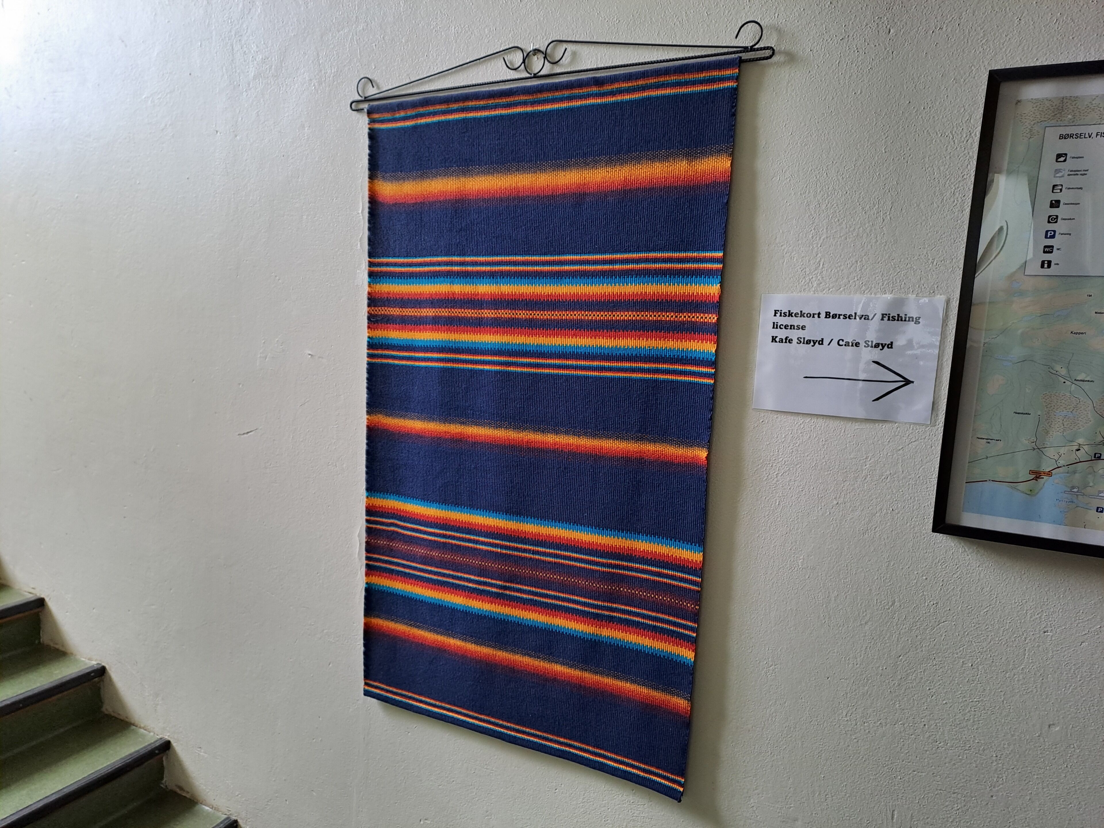 A vertical textile hanging on a wall, a directional sign, and a framed map.