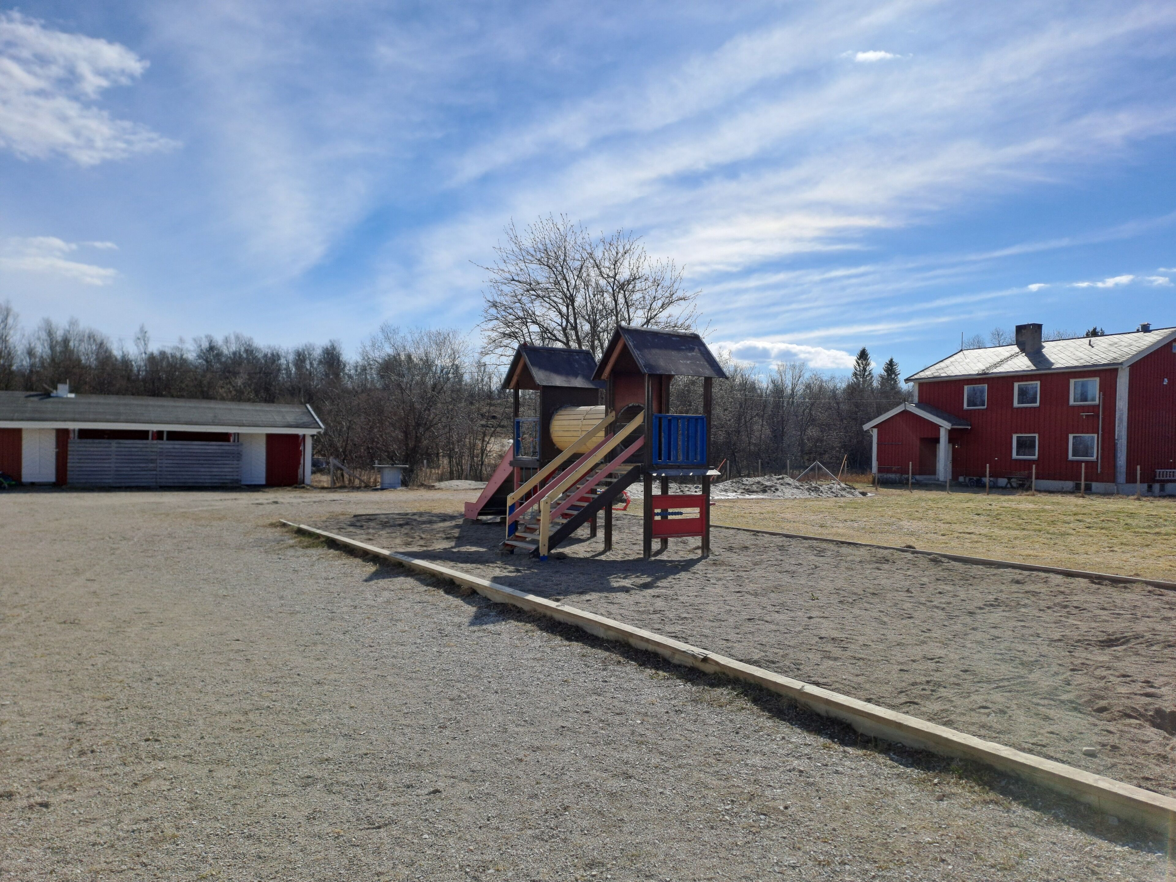 Une aire de jeux avec un toboggan et des escaliers, un bâtiment rouge et une zone recouverte de gravier.
