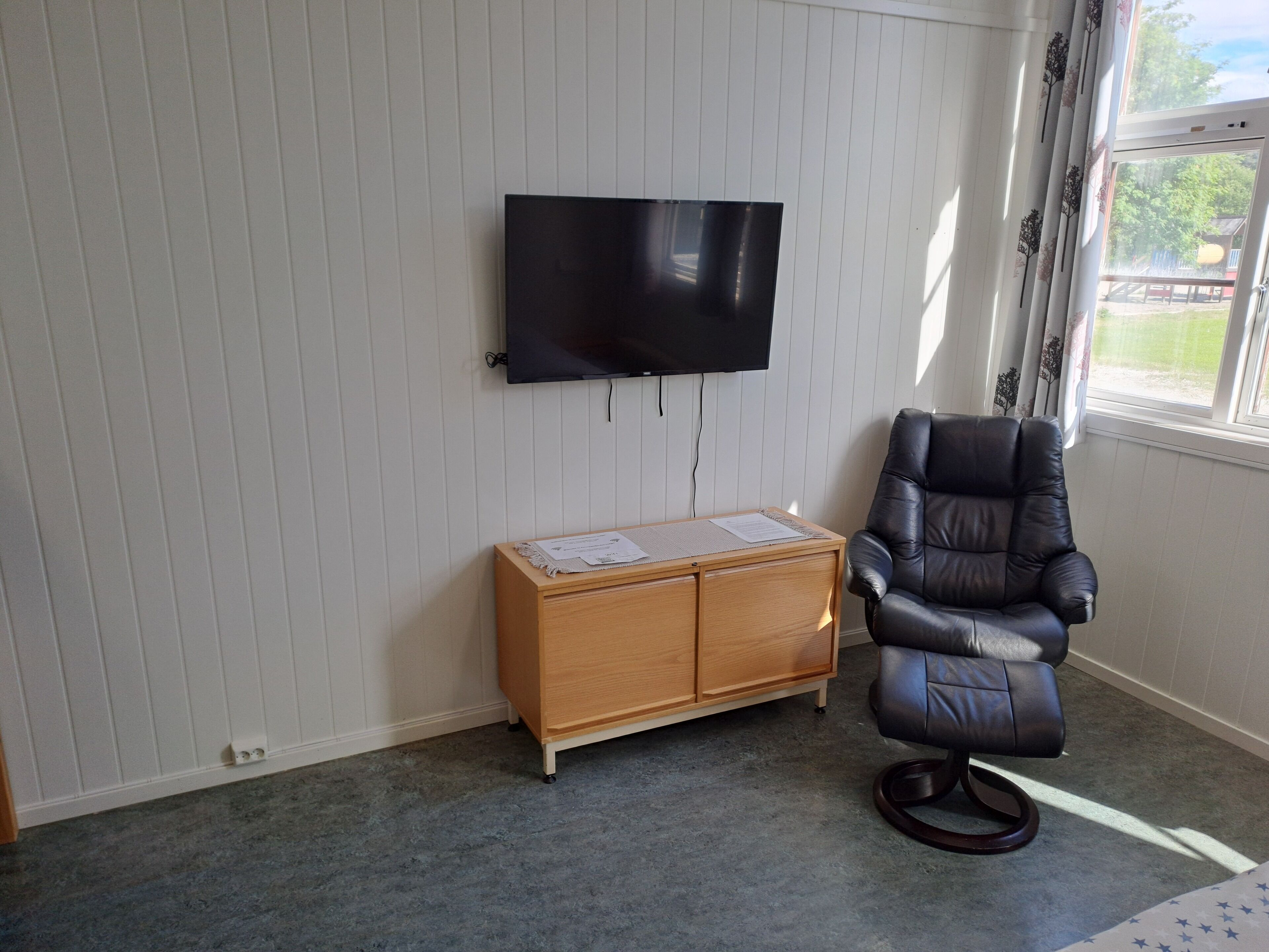 A room with a flat-screen TV mounted on the wall, a wooden cabinet with papers, a leather chair, and a window with curtains.