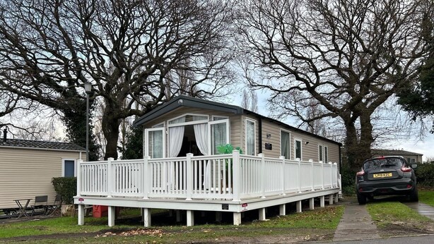 A single-story mobile home with a white porch and a small deck, surrounded by trees. - Rockley Park (Poole)