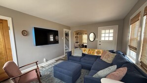 Ein Wohnzimmer mit einem blauen Ecksofa, einem Schaukelstuhl, einem an der Wand montierten Flachbildfernseher und einem gemusterten Teppich.
