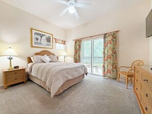 Ein Schlafzimmer mit einem Bett, zwei Nachttischen, einer Kommode, einem Deckenventilator, einem Fenster mit Vorhängen und einem gerahmten Bild an der Wand.