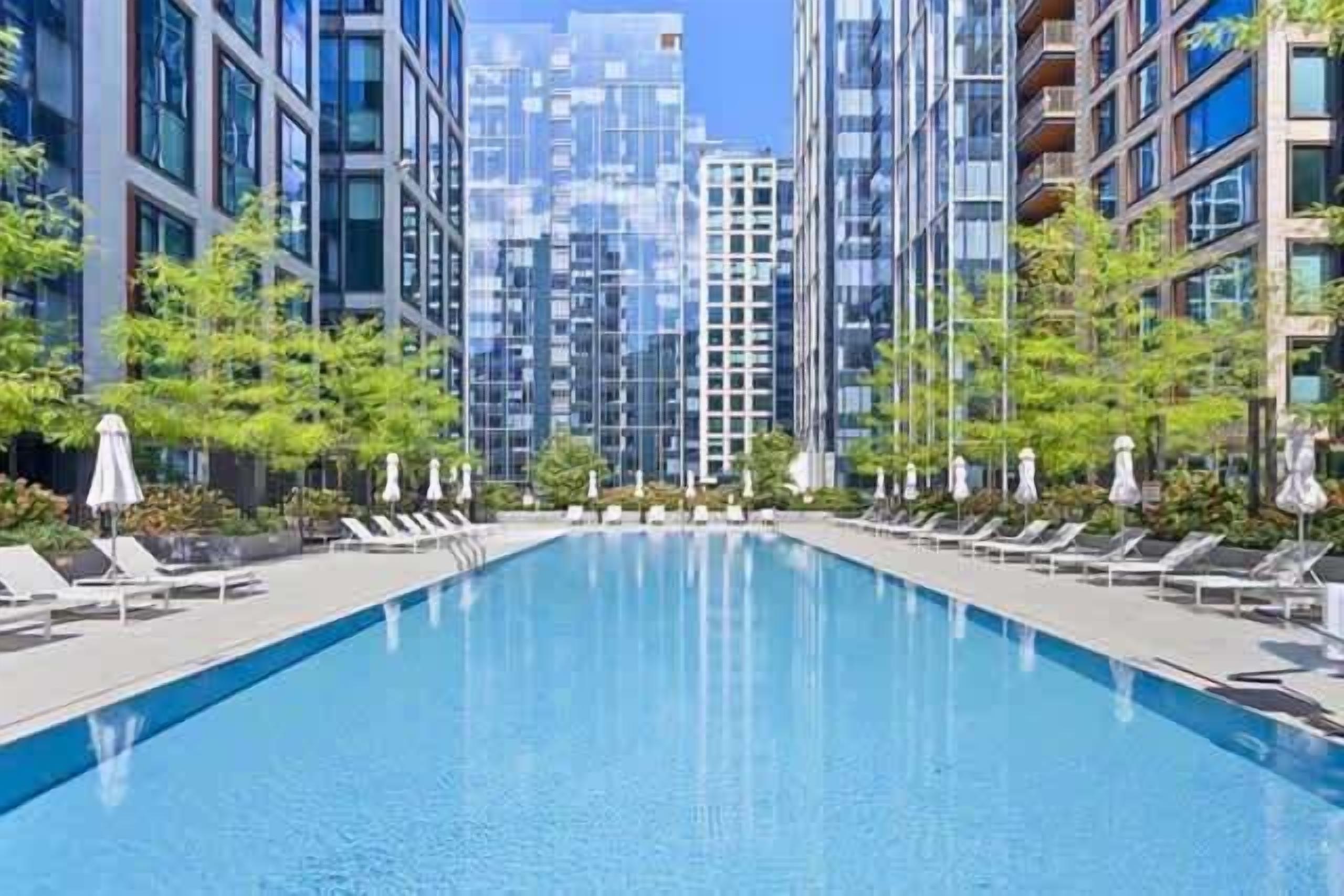 A swimming pool with lounge chairs and umbrellas, surrounded by modern buildings.