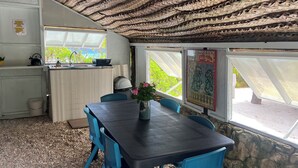 A room with a woven ceiling, a table with chairs, a window with a view, and a kitchen area with a kettle and a bowl. - Fare Miki Miki (Huahine)