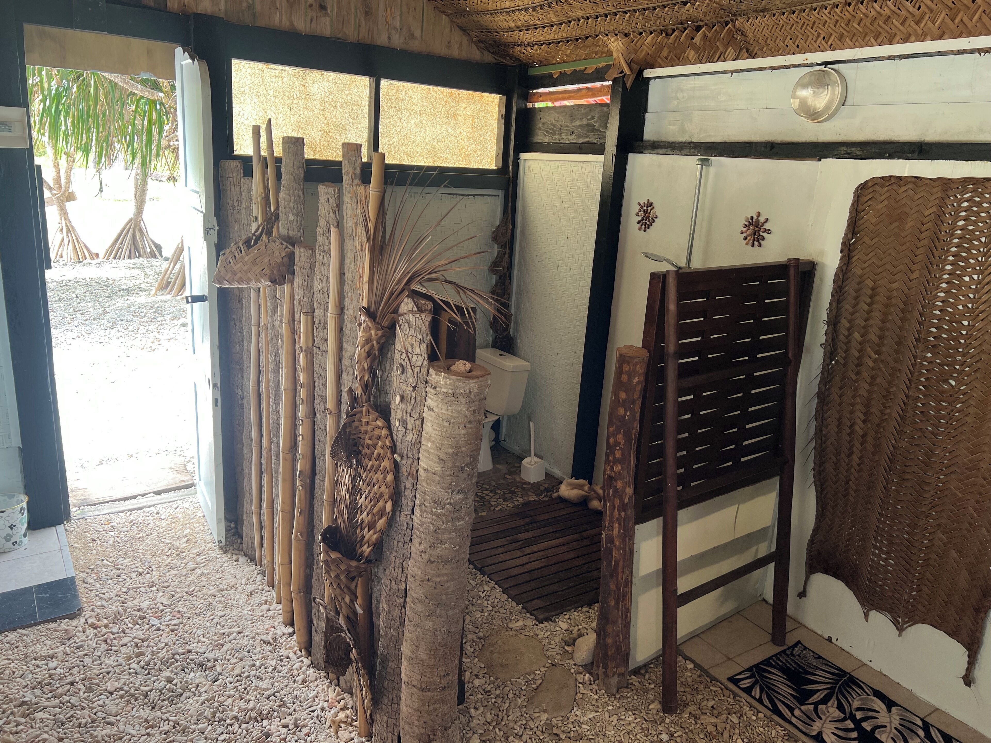 A room with a thatched roof, a toilet with a bamboo shower curtain, and a woven basket hanging on the wall.