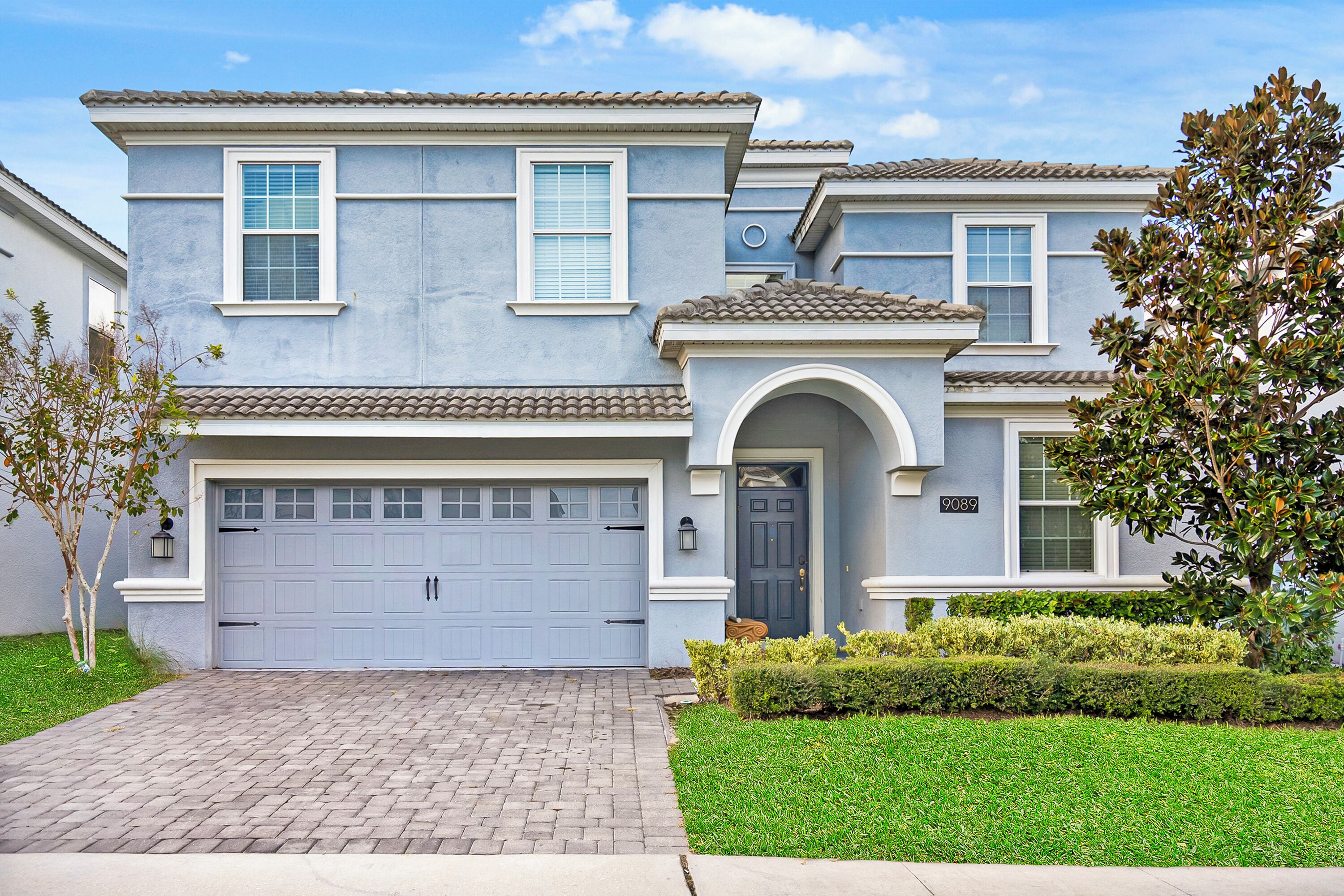 A two-story residential building with a garage, a front door, and a small front yard with a tree and shrubs.