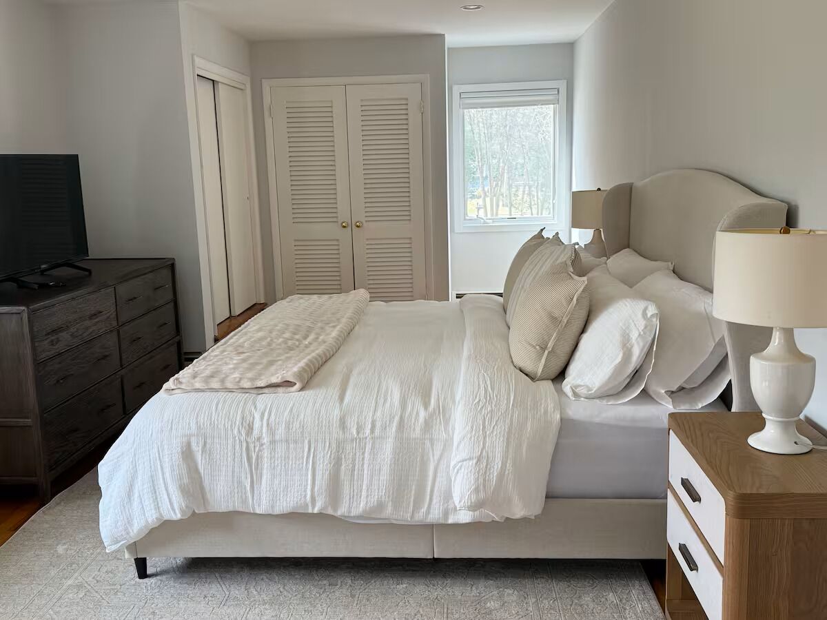 Une chambre à coucher comprenant un lit, une commode, une table de chevet, une lampe et une fenêtre donnant sur un beau paysage.