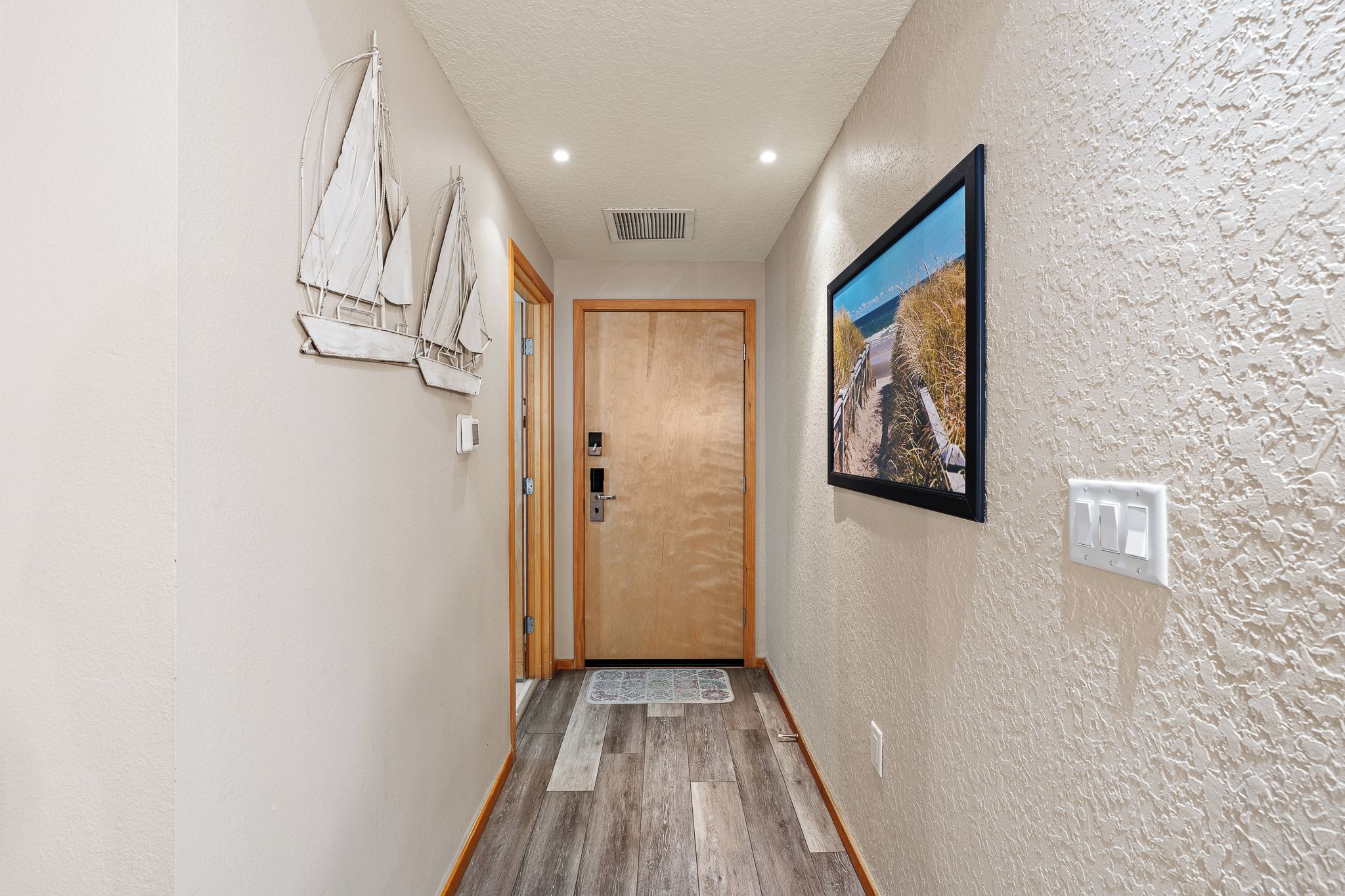 Un pasillo con una puerta de madera, un cuadro enmarcado de una playa e interruptor de luz en la pared.