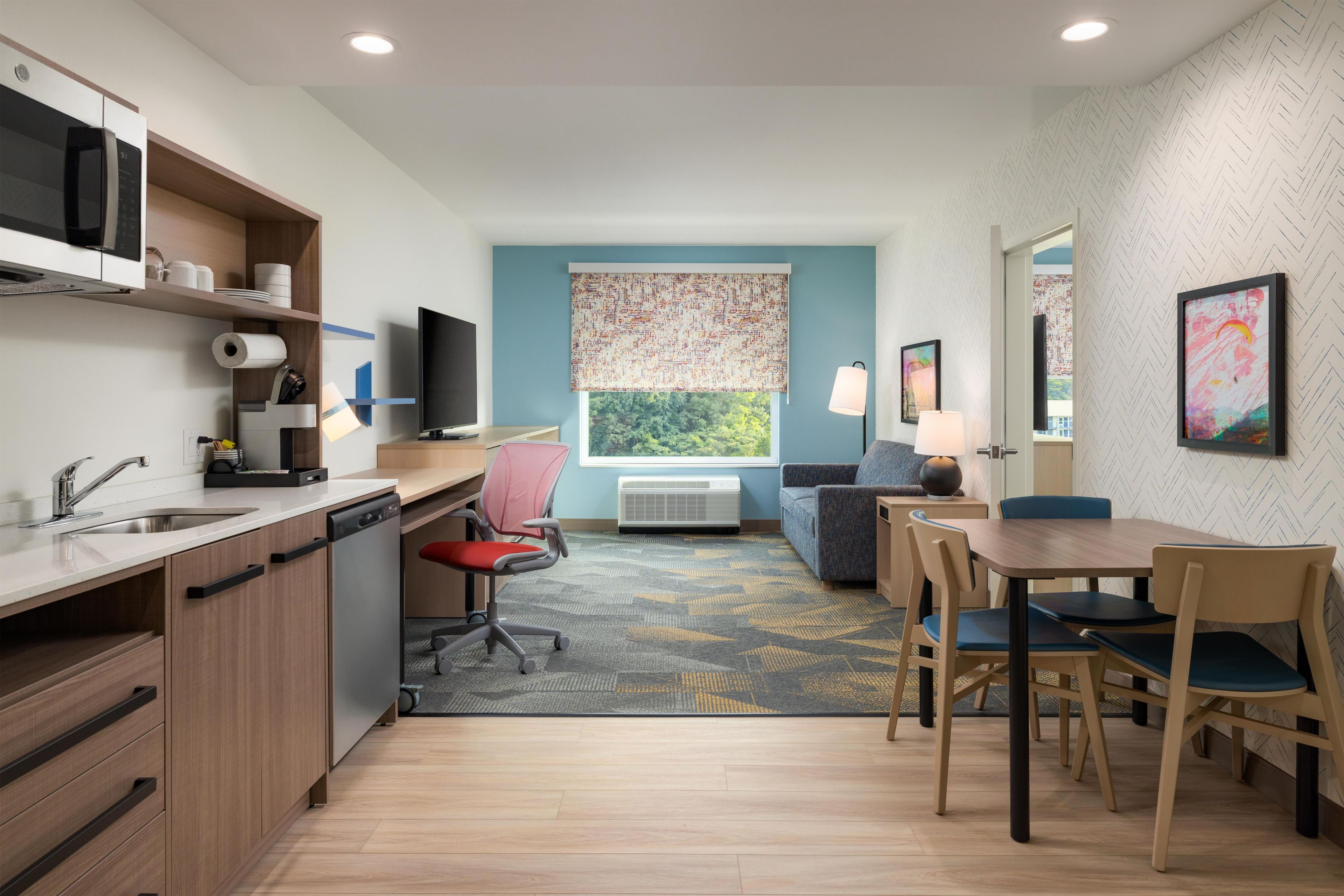 A compact hotel room with a kitchenette, a dining area with a table and chairs, a sofa, a TV, and a large window with a view.