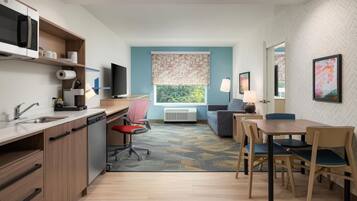A compact hotel room with a kitchenette, a dining area with a table and chairs, a sofa, a TV, and a large window with a view.