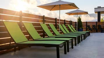 A row of green lounge chairs with umbrellas, arranged against a wooden fence, under a clear sky.