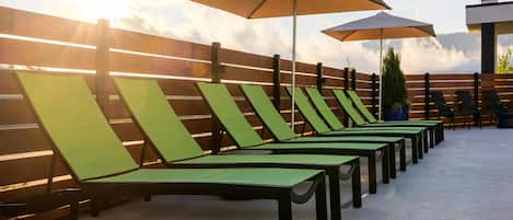 A row of green lounge chairs with umbrellas, arranged against a wooden fence, under a clear sky.
