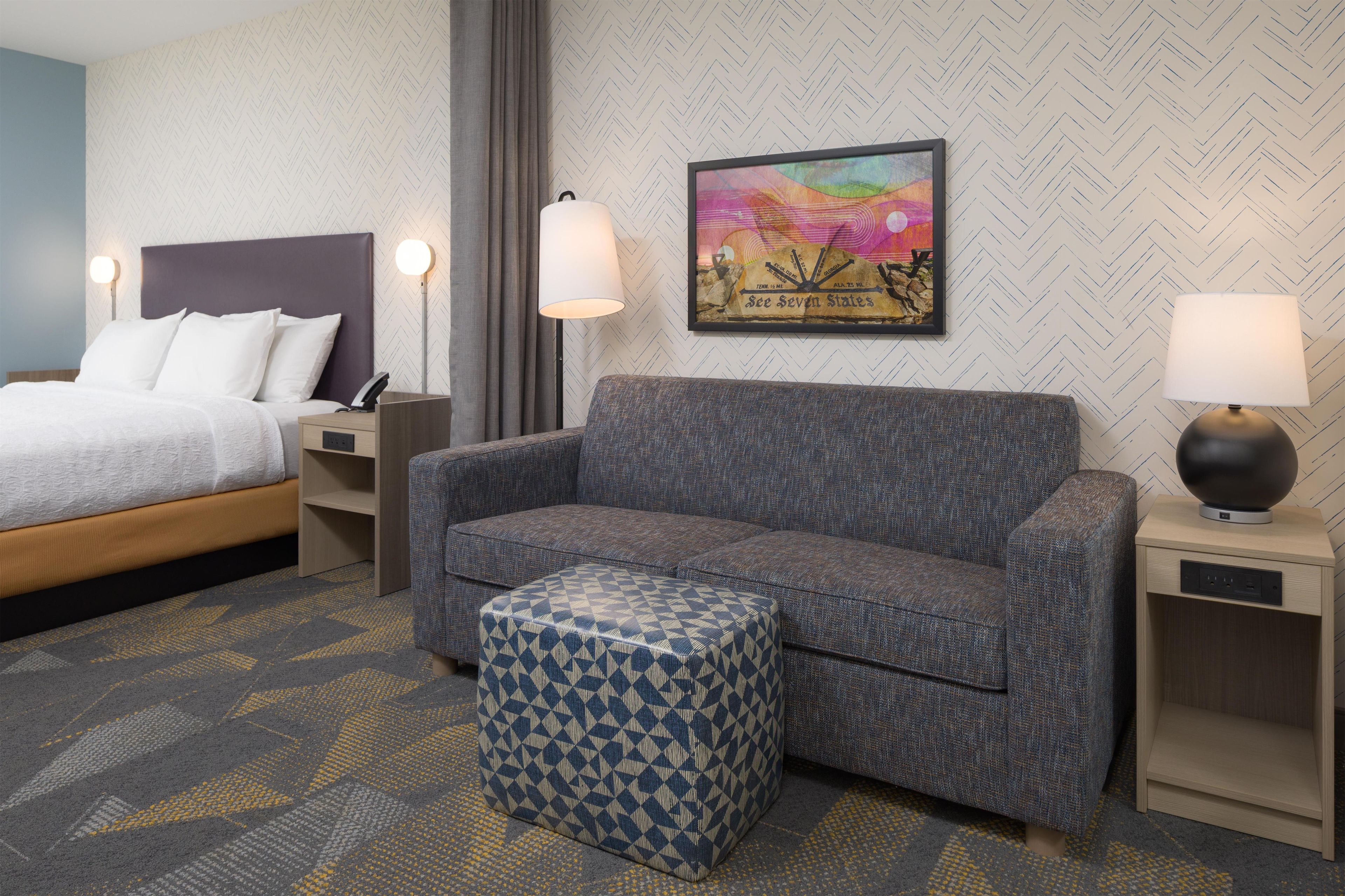 Habitación de hotel con cama, sofá, reposapiés, mesita de noche, lámpara y pared con patrón de espiga.