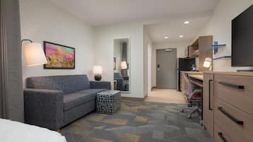 A hotel room with a sofa, a small ottoman, a desk with a chair, a television, and a bathroom visible through an open door.