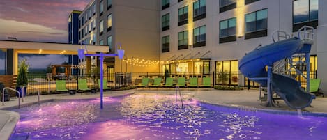 A hotel pool area with a water slide, lounge chairs, and a view of the building.