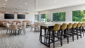 A modern dining area with a long table and multiple chairs, large windows with a view of greenery, and a flat-screen TV mounted on the wall.