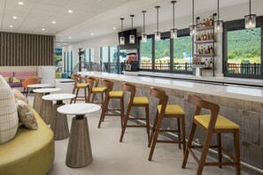 Eine Bar mit mehreren Hockern und einer Theke, ein Sitzbereich mit runden Tischen und Sofas sowie ein Blick ins Grüne durch große Fenster.