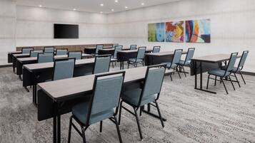 A conference room with multiple tables and chairs, a flat-screen TV mounted on the wall, and abstract art on the back wall.