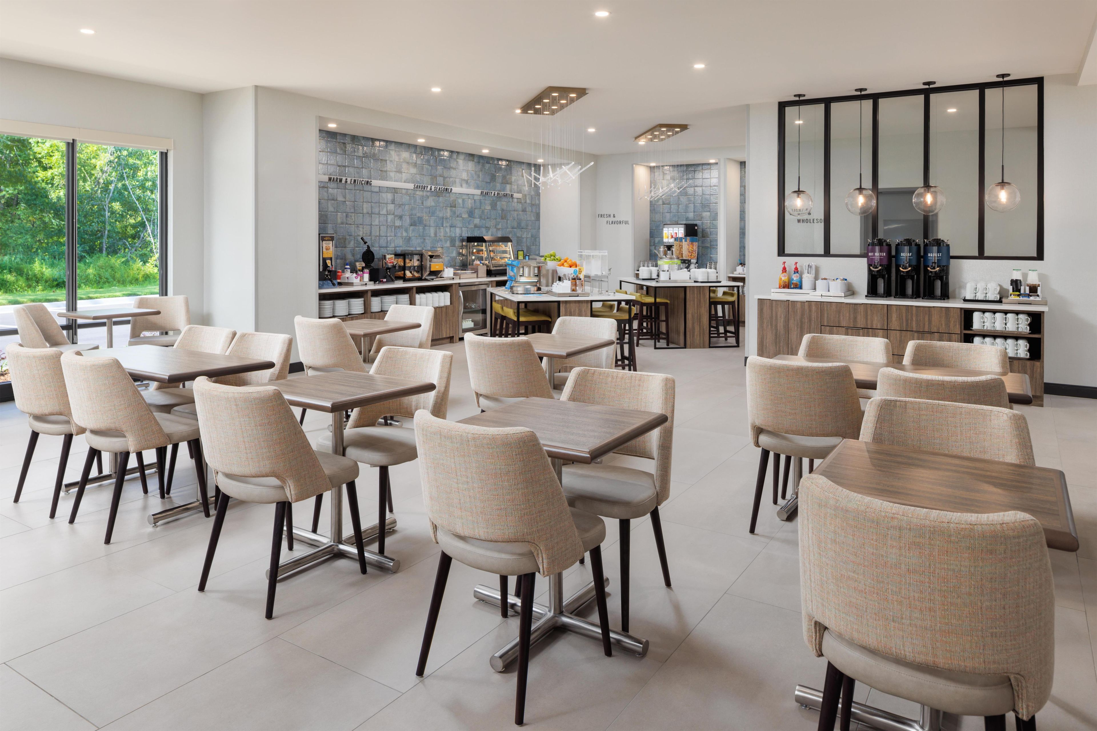 A modern dining area with multiple tables and chairs, a kitchen area with a coffee station, and large windows offering a view of greenery.
