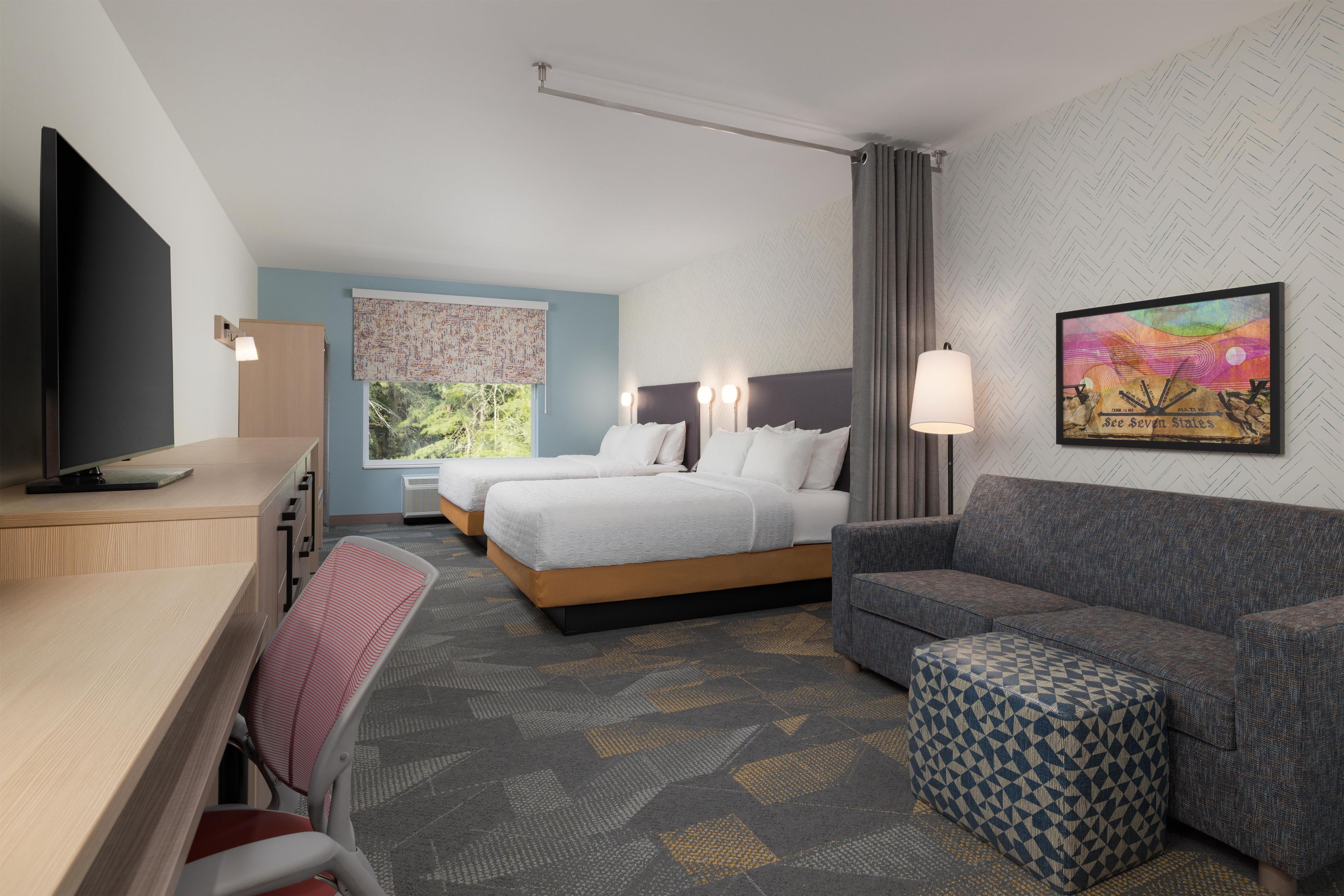 A hotel room with a bed, a desk, a chair, a sofa, and a window with a view of greenery.