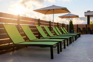 Une rangée de fauteuils de jardin verts, dotés de parasols, est disposée le long d’une clôture en bois, sous un ciel dégagé.