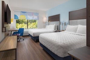 A hotel room with two beds, a desk, a chair, and a window with a view of trees.