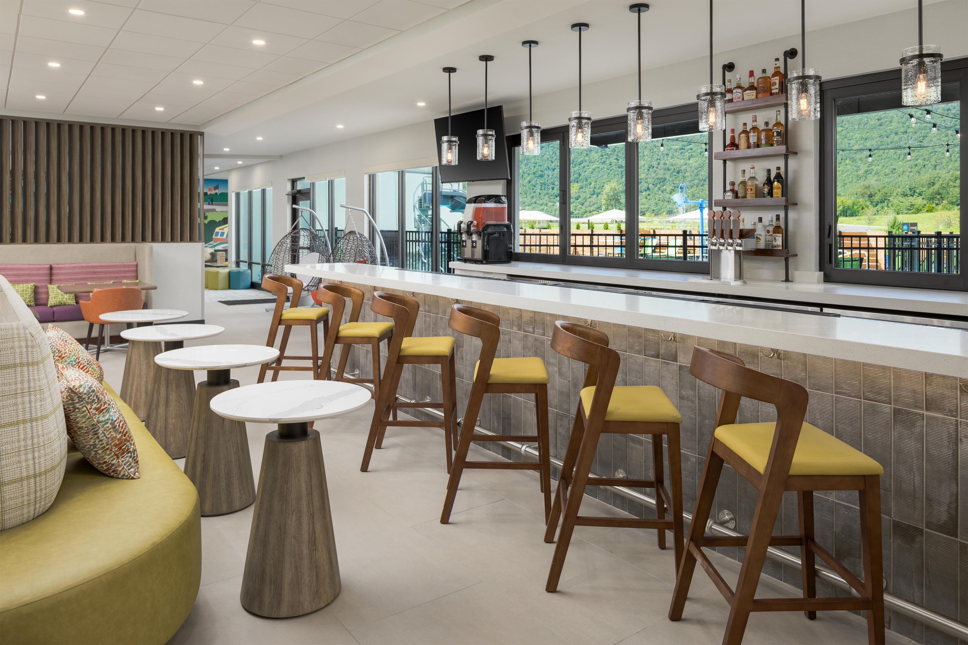 A bar area with multiple stools and a counter, a seating area with round tables and sofas, and a view of greenery through large windows.