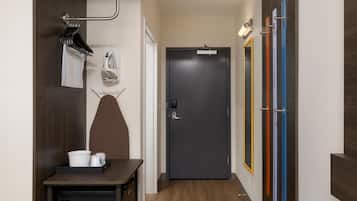 A hallway with a dark door, a coat rack with hangers, a small table with a basket, and a wall-mounted ironing board.