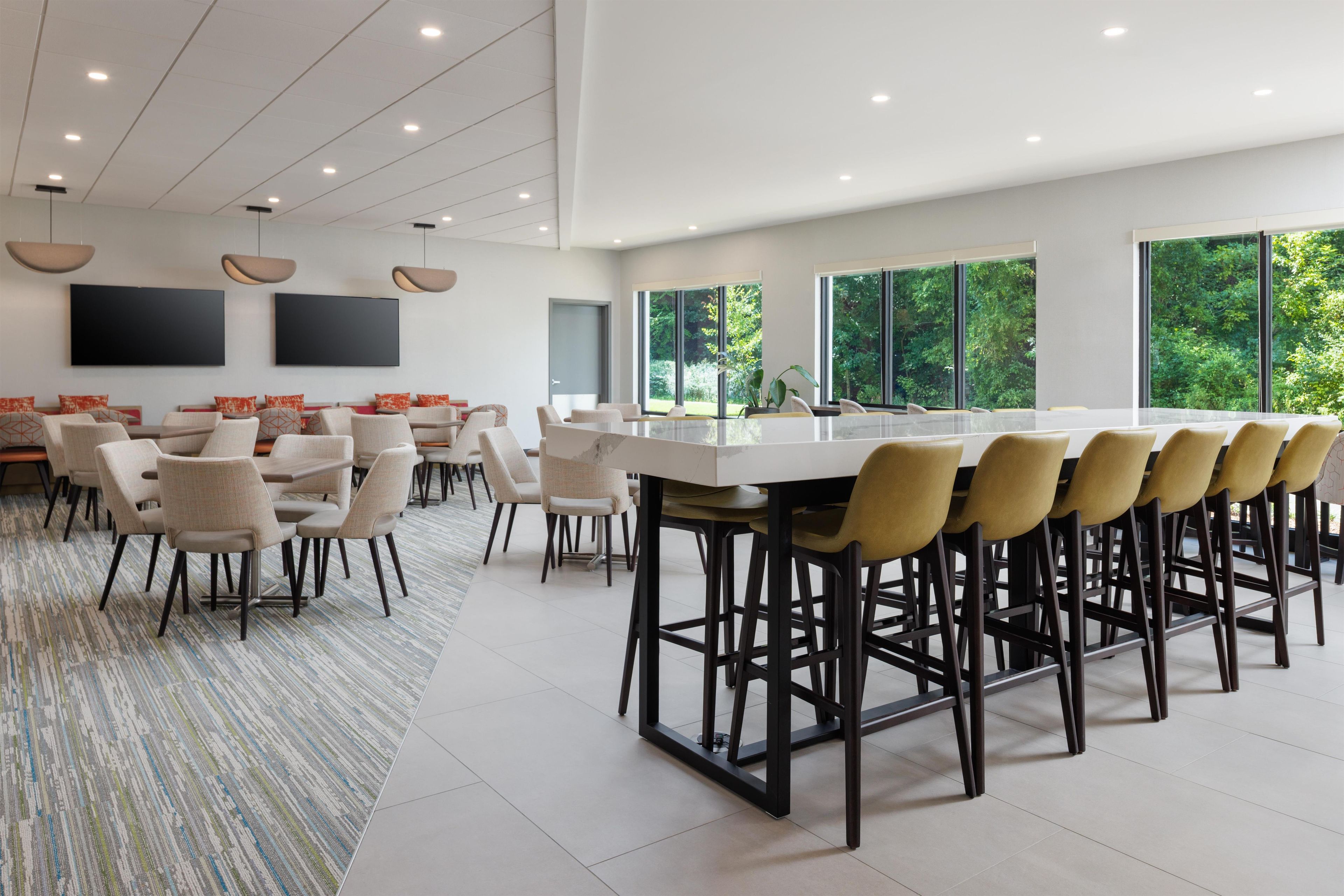 A modern dining area with a long table and multiple chairs, large windows with a view of greenery, and a flat-screen TV mounted on the wall.