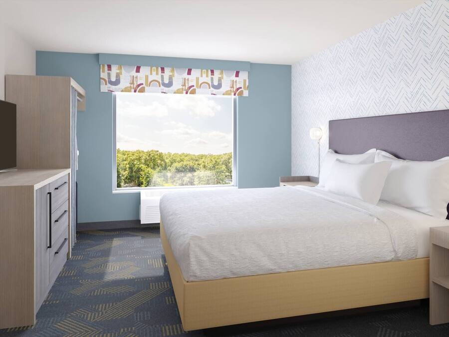 A hotel room with a bed, a TV, a desk, and a window with a view of trees.
