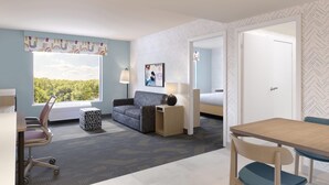 Quarto de hotel com cama, escrivaninha, poltrona, sofá, luminária e janela com vista para árvores.