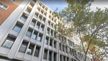 Un edificio de varios pisos con fachada de diseño geométrico, grandes ventanales y un árbol en primer plano.