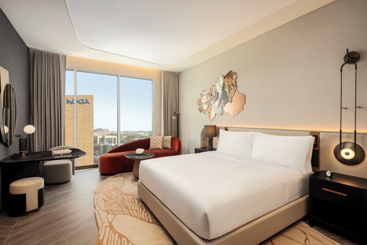 Habitació d'hotel moderna amb un llit gran, un sofà vermell, una taula petita i vistes a l'horitzó urbà.