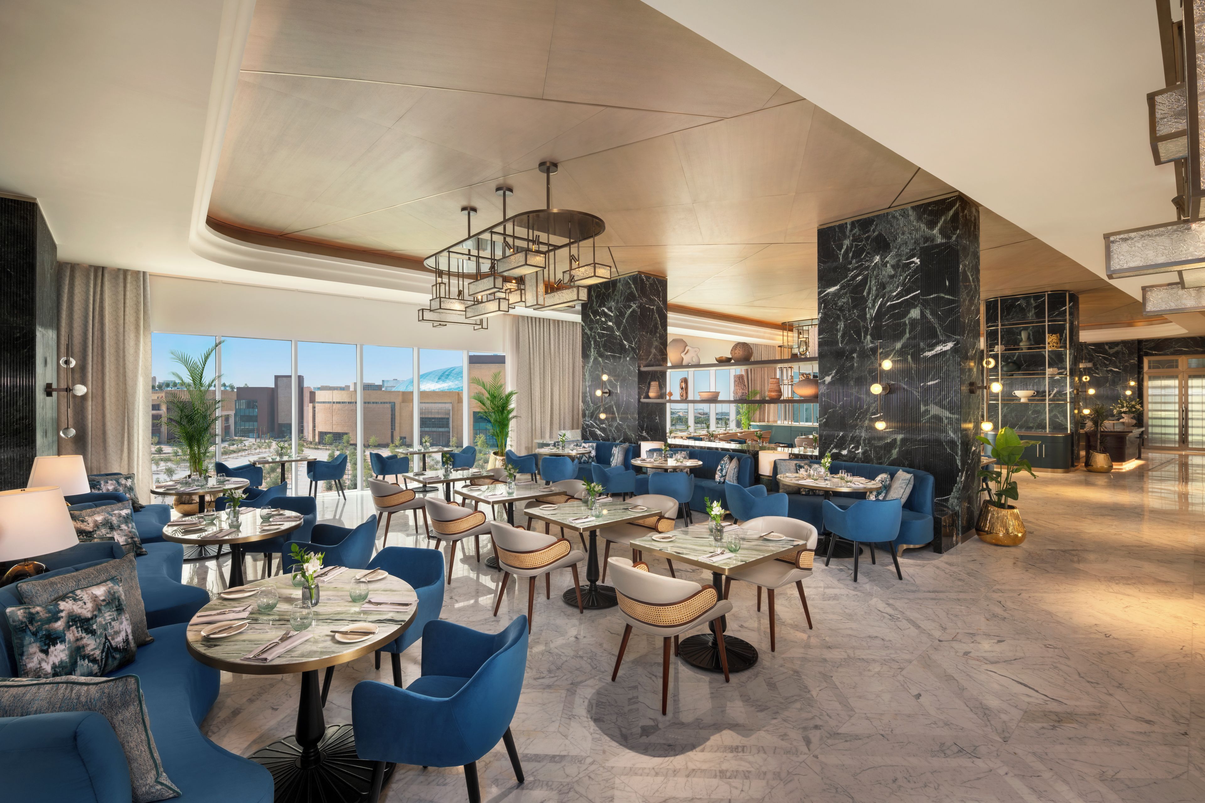 A modern restaurant interior with round tables, blue chairs, and a view of the cityscape through large windows.