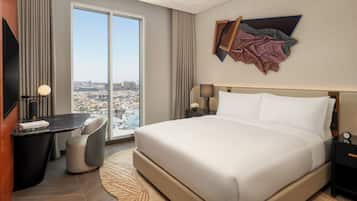 A hotel room with a large bed, a desk, a chair, a nightstand, a lamp, and a view of the city.