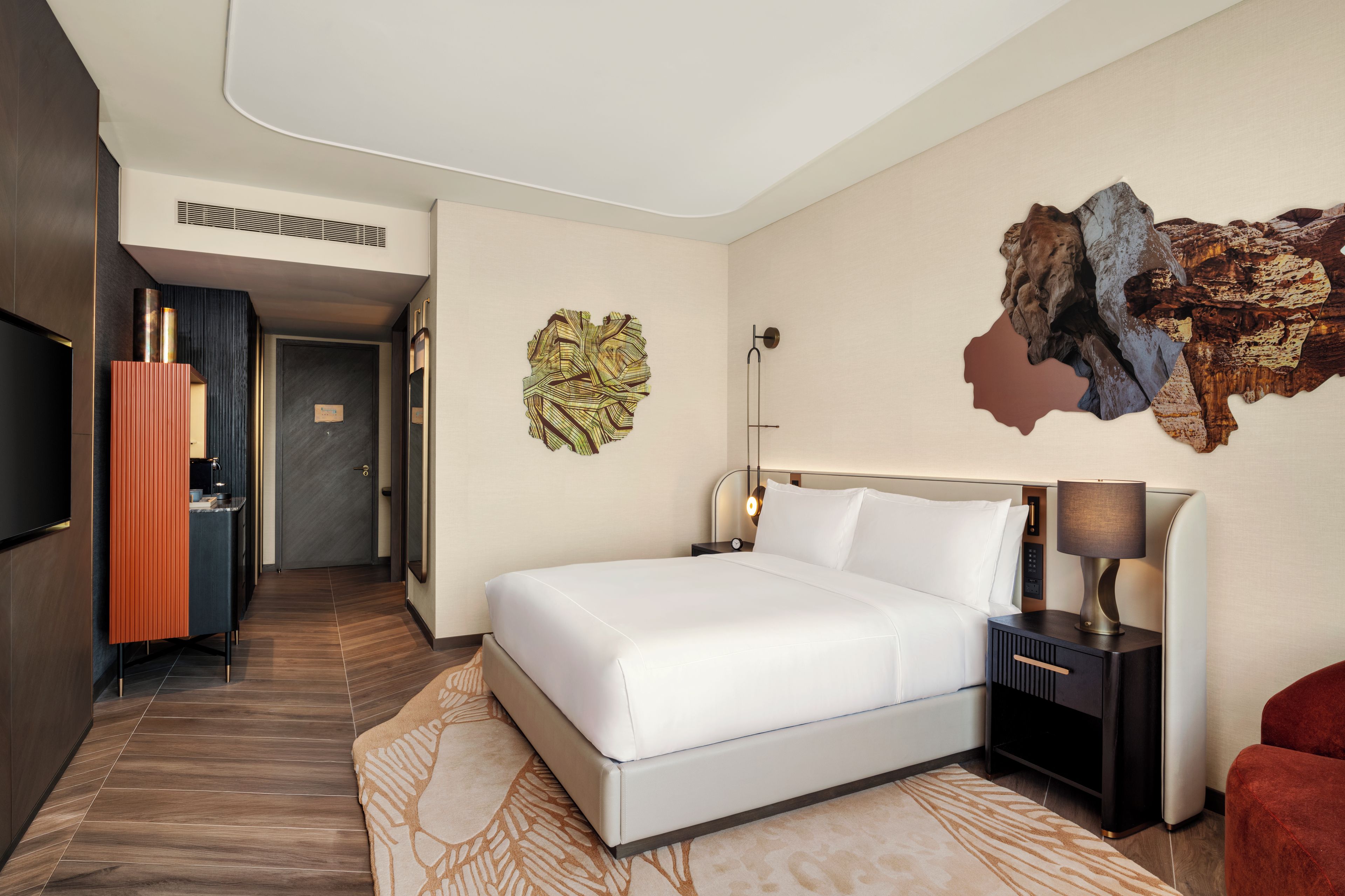 A hotel room with a bed, a nightstand, a lamp, a red chair, and a wall art piece.