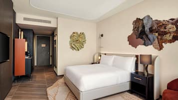 A hotel room with a bed, a nightstand, a lamp, a red chair, and a wall art piece.