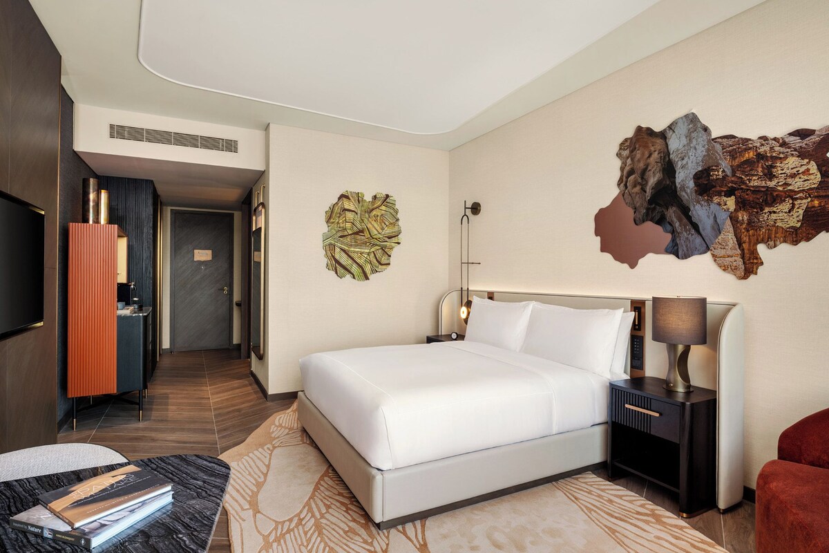 Habitació d'hotel amb un llit, una tauleta de nit, una làmpada, una cadira vermella, una obra d'art a la paret i una televisió.
