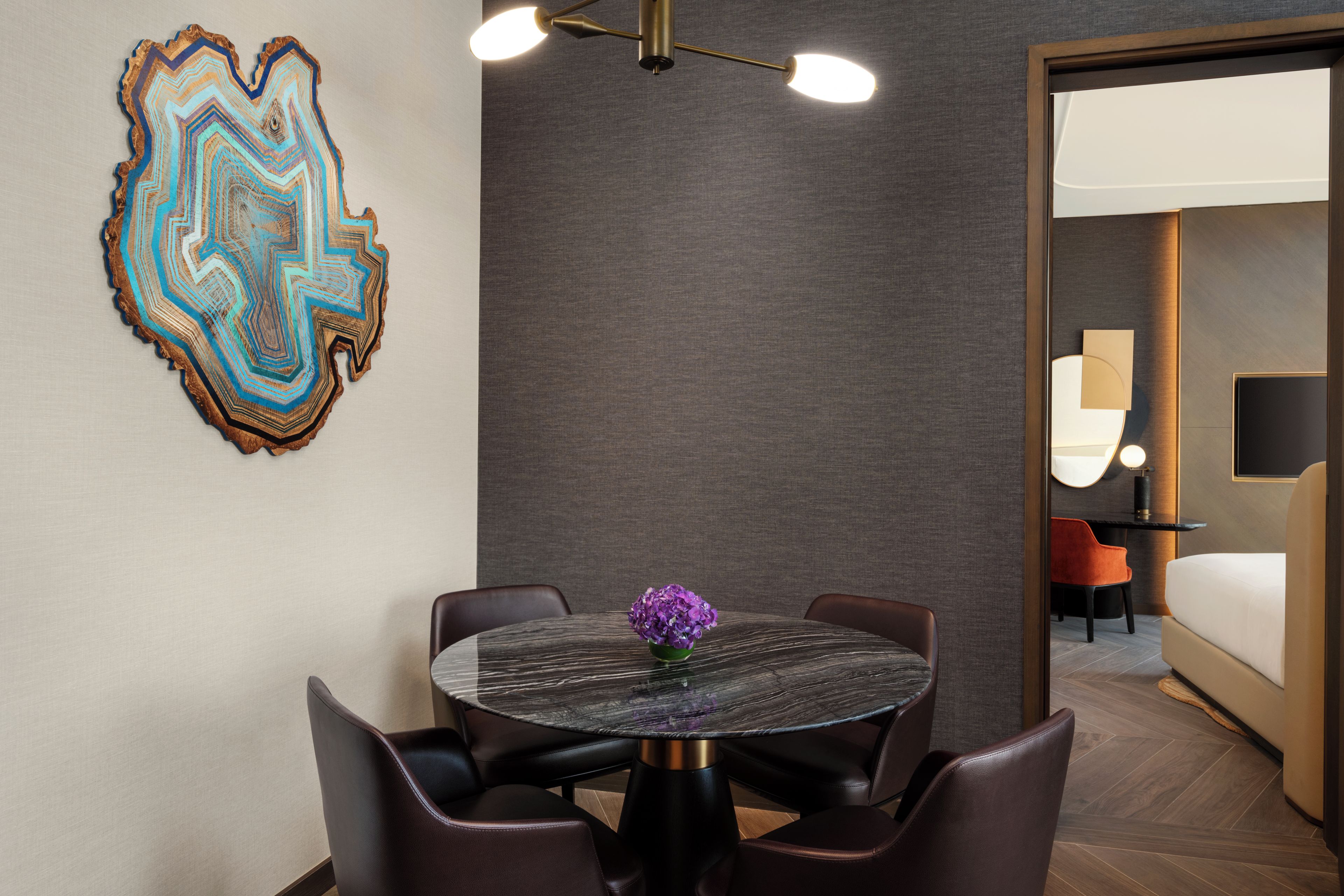 A modern hotel room with a round dining table, leather chairs, a decorative wall piece, and a view into the adjacent room.